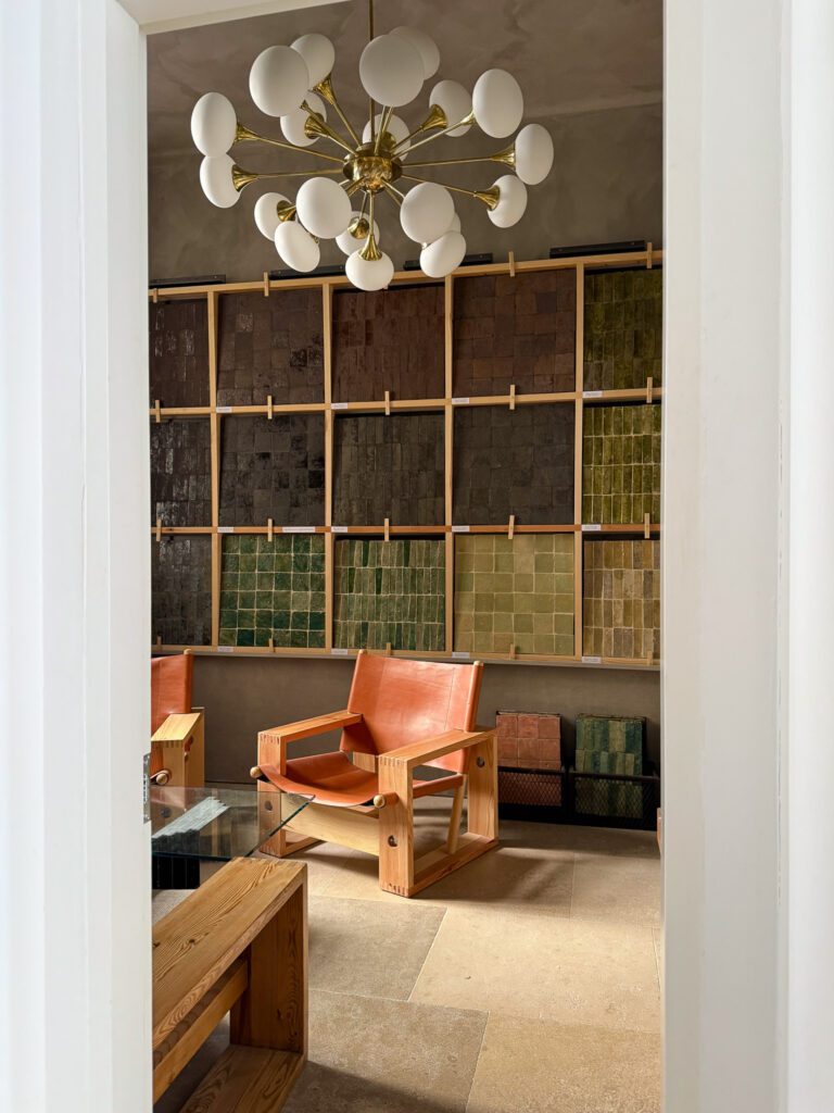 Ett modernt rum med en trästol och ett bord i förgrunden, en vägg med färgglada kakelprover och en stor vit och mässingsfärgad ljuskrona ovanför.