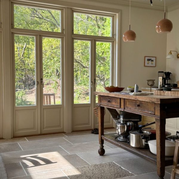 Ljust kök, antikbehandlat kalkstensgolv, rustik köksö i trä, hängande lampor och fönsterutsikt mot träd. Varm och inbjudande atmosfär.