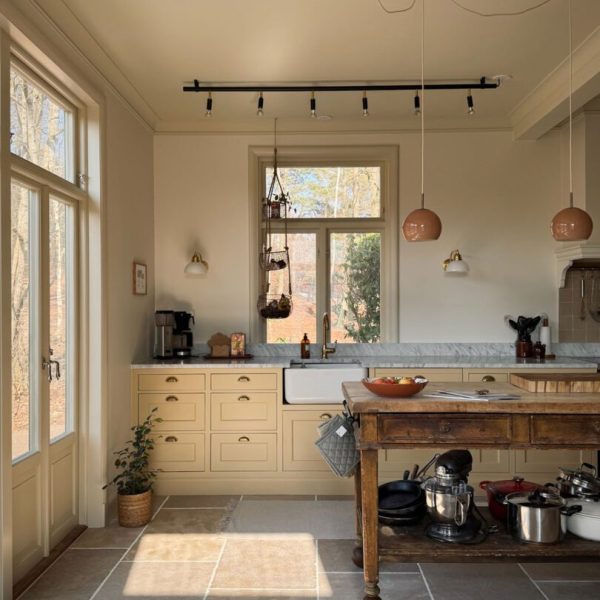 Ljust kök med gula skåpsluckor, antikbehandlat kalkstensgolv, rustik köksö i trä, hängande lampor och fönsterutsikt mot träd. Varm och inbjudande atmosfär.