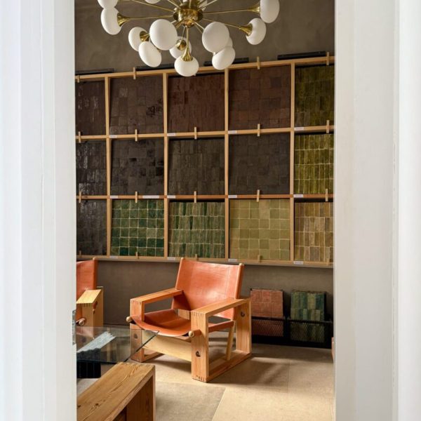 Ett modernt rum med en trästol och ett bord i förgrunden, en vägg med färgglada kakelprover och en stor vit och mässingsfärgad ljuskrona ovanför.